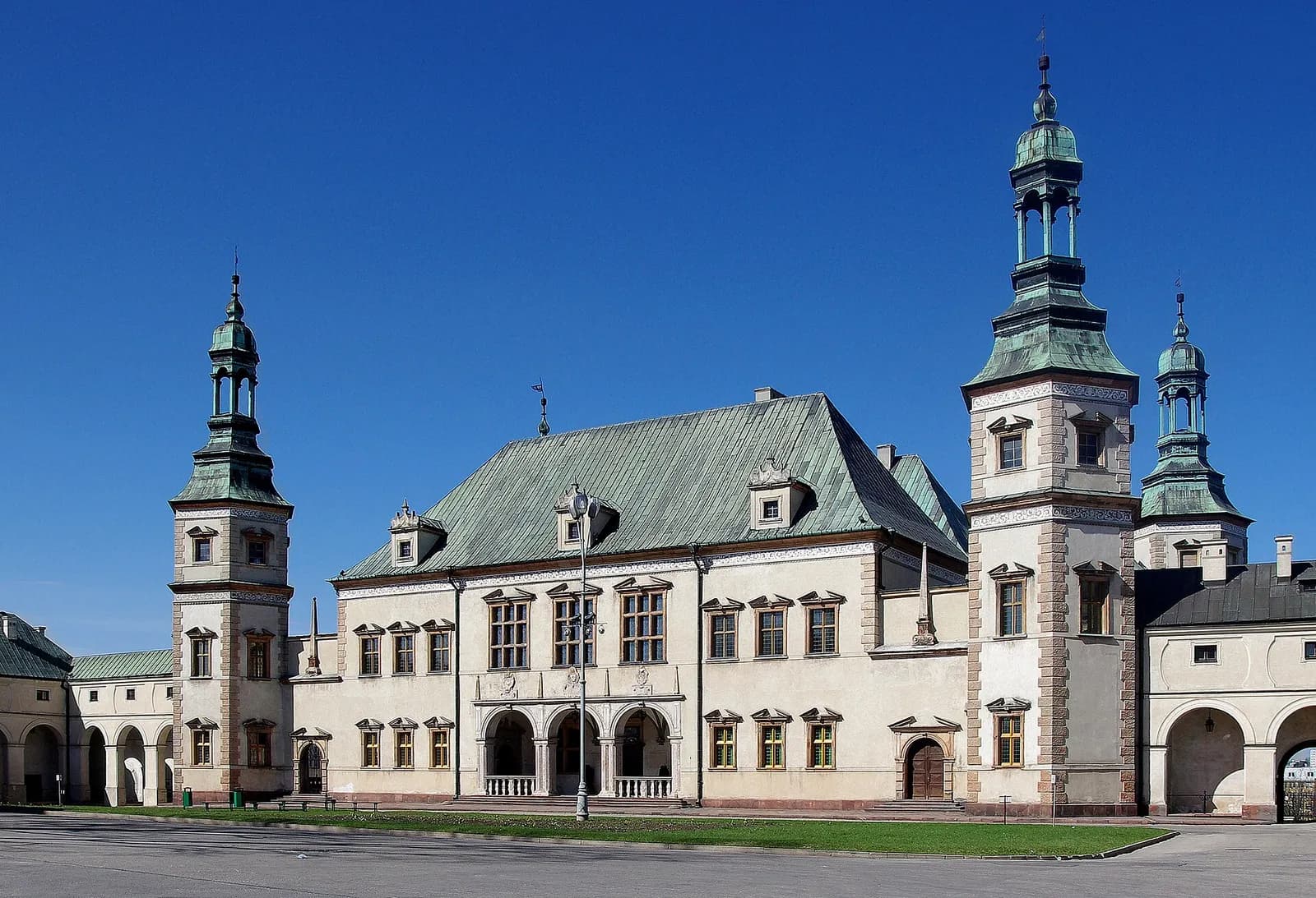 A historic, light-colored building with a large peaked roof and two prominent towers with green, ornate spires stands against a clear blue sky, with an arched walkway visible on the left.