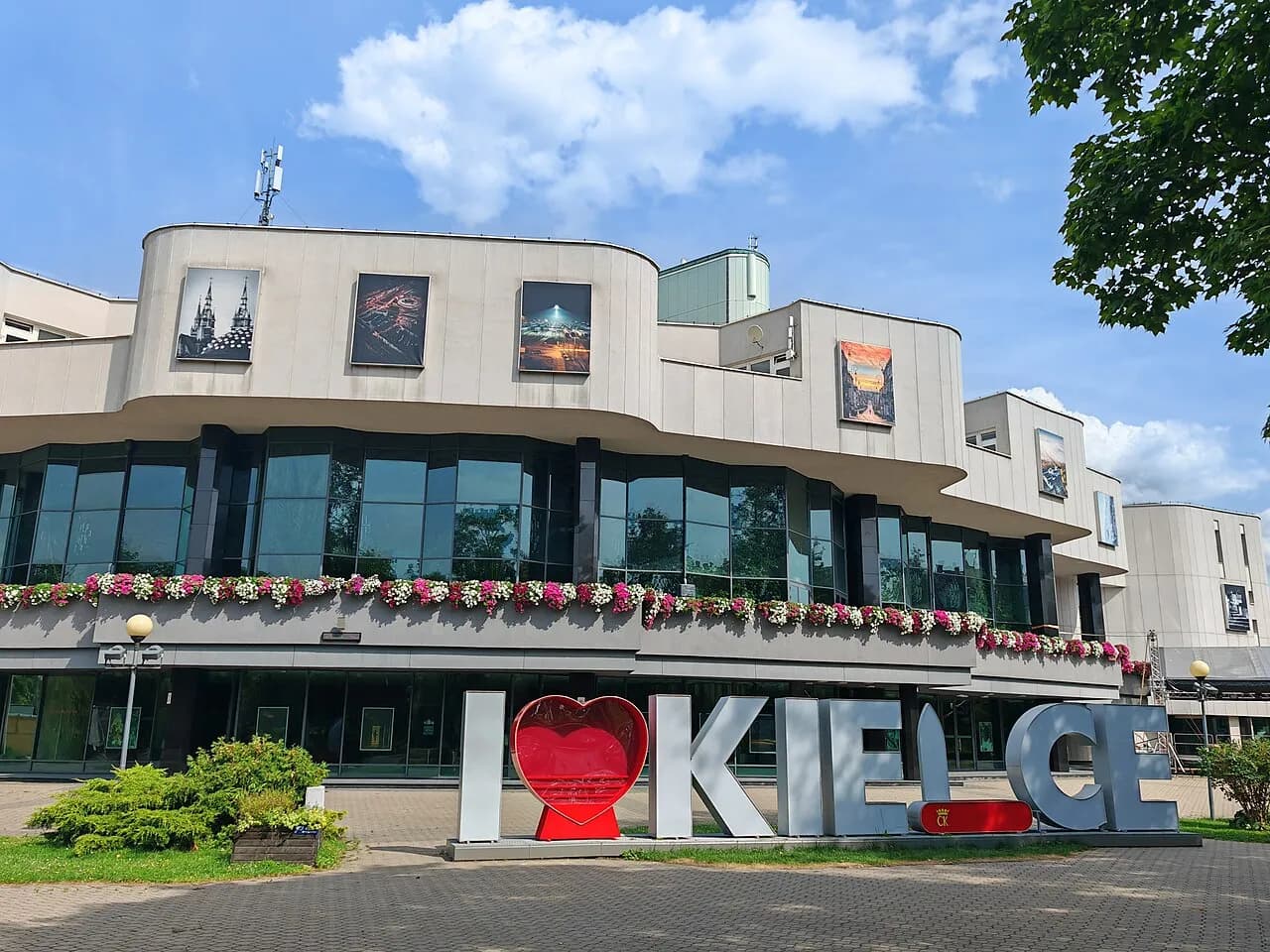 A modern building with a curved facade and large windows, adorned with flower boxes, stands behind large letters on the ground spelling out "I β€οΈ KIELCE."